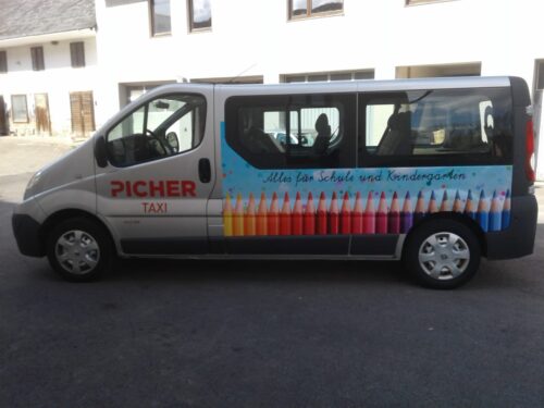 Seitliche Ansicht eines weißen Kleinbusses mit der Aufschrift 'PICHER TAXI' und einer bunten Buntstiftgrafik sowie dem Schriftzug 'Alles für Schule und Kindergarten'
