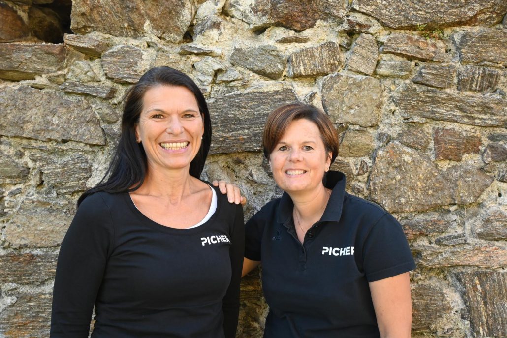 Zwei Frauen in schwarzen Shirts mit der Aufschrift 'PICHER' vor einer grob behauenen Steinmauer, eine legt die Hand auf die Schulter der anderen